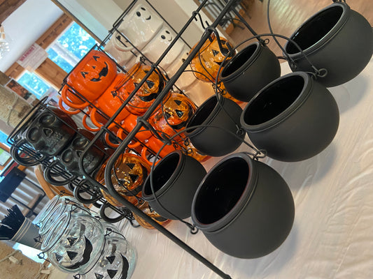 A display of Halloween-themed products including matte black hanging cauldrons, orange jack-o'-lantern lanterns, and clear glass jars with black jack-o'-lantern cutouts, arranged on black metal racks over a white tablecloth.