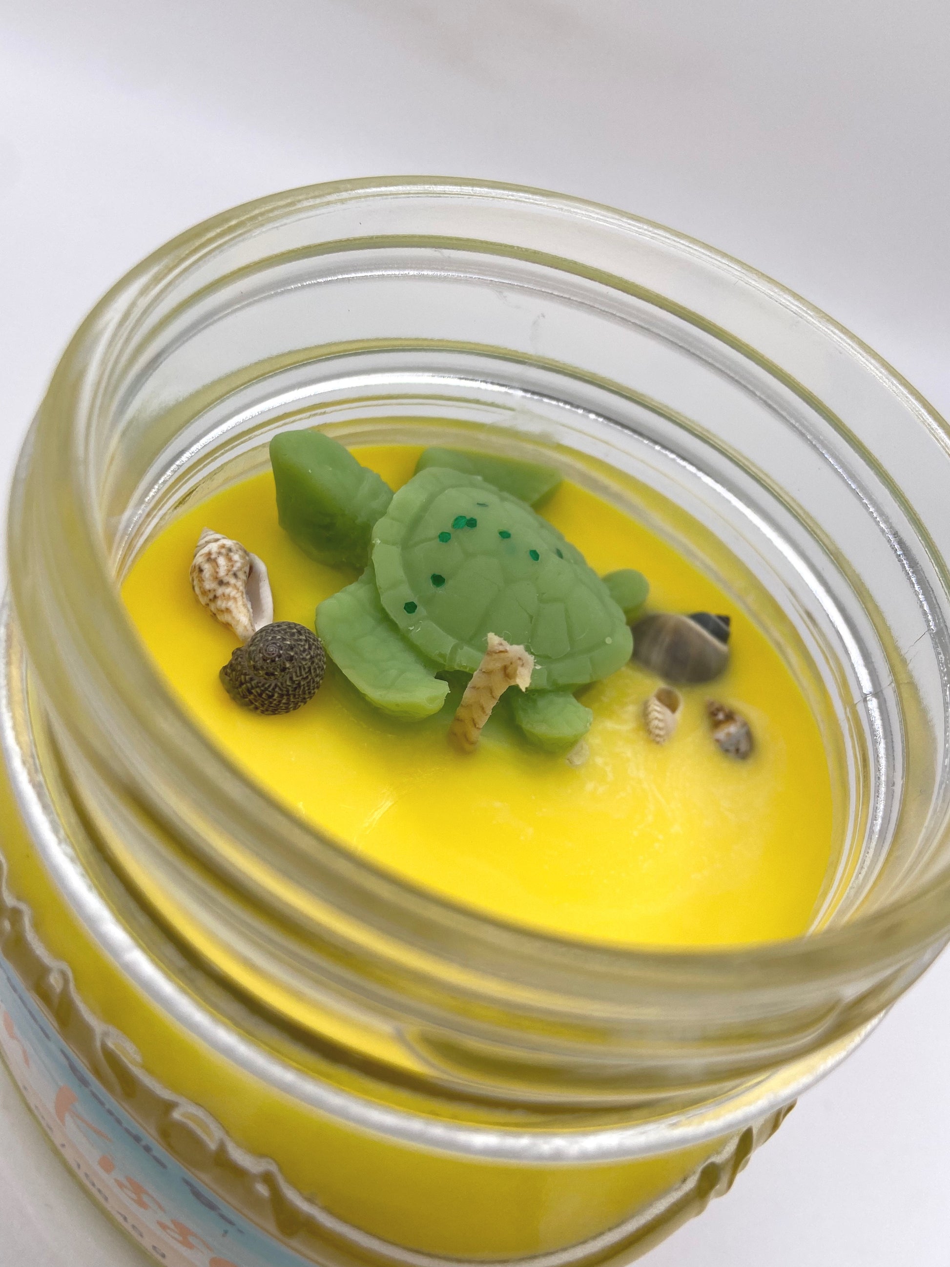 Jar with a yellow candle and green turtle-shaped wick trimmer on a white background